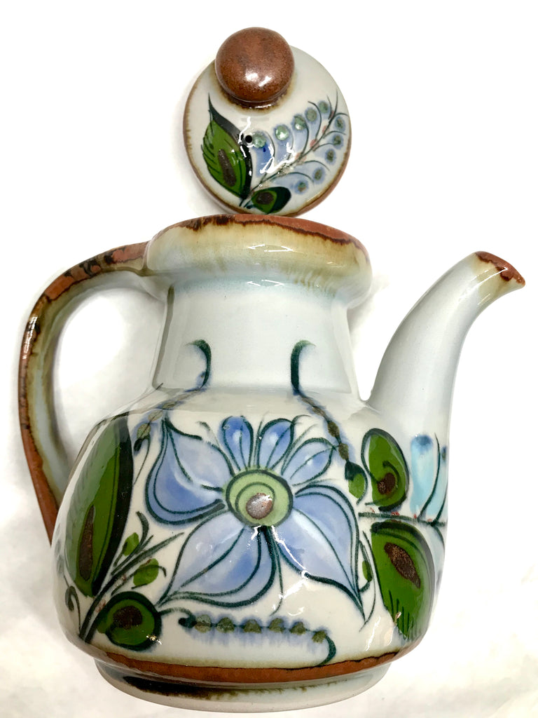 A teapot with a lid with brown trim and grey background with green leaves and blue flowers.