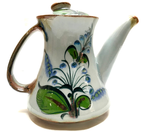 Coffee pot with lid and handle brown trim, grey clay background green leaves and blue flowers.