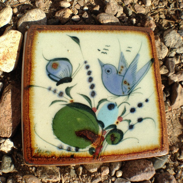 Ken Edwards tile with brown rim.   It is natural grey clay color background with birds, butterflies, and leaves in blue, green, black and brown on the face.