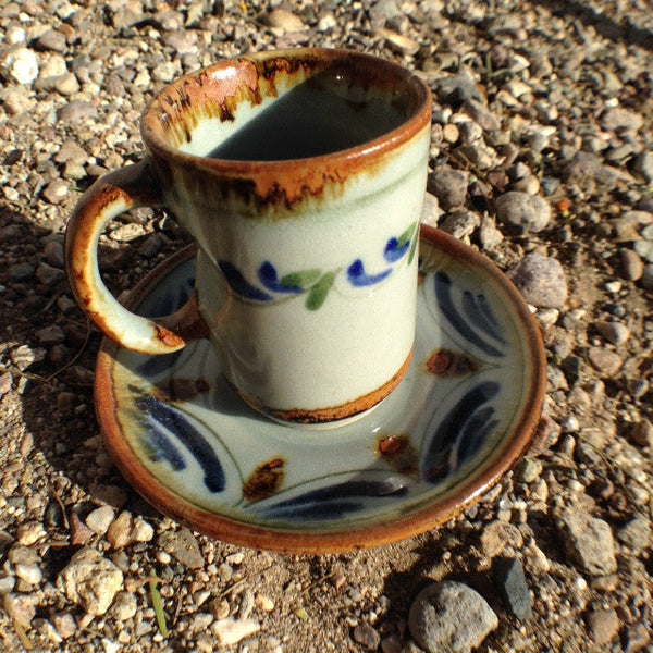 Ken Edwards Expresso cup and saucer set.  Brown rim on both pieces.  It is natural grey clay color background with birds, butterflies, and leaves in blue, green, black and brown on the outside. 