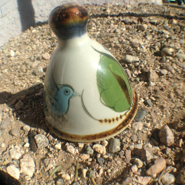 This Ken Edwards stoneware bell has a brown rim and handle.   It is natural grey clay color background with birds, butterflies, and leaves in blue, green, black and brown on the outside.